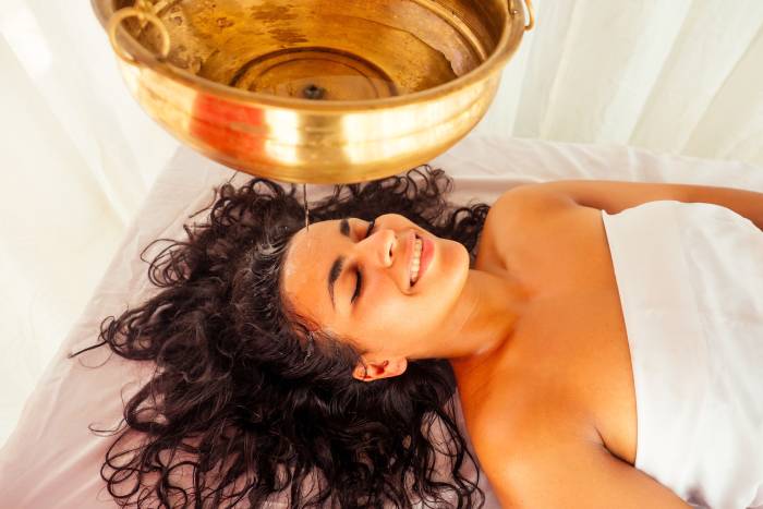 Image of a woman lying on a white bed with open hairs and a white cloth wrapped around with a pan filled with oil hanging above her head getting  ayurvedic "shirodhara"  treatment.